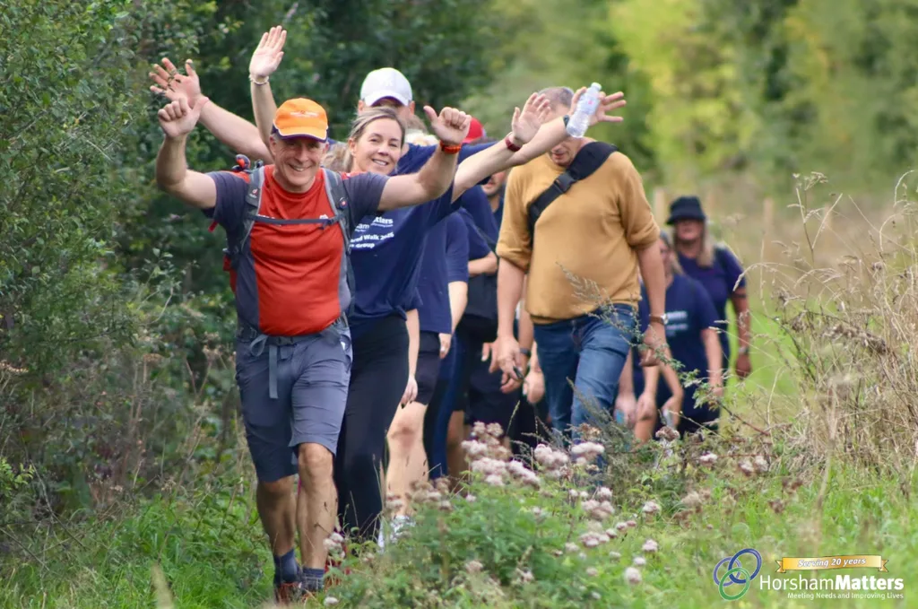 Walkers in Horsham Matters T-shirts wave as they walk single file along a grassy path on the 20km sponsored walk